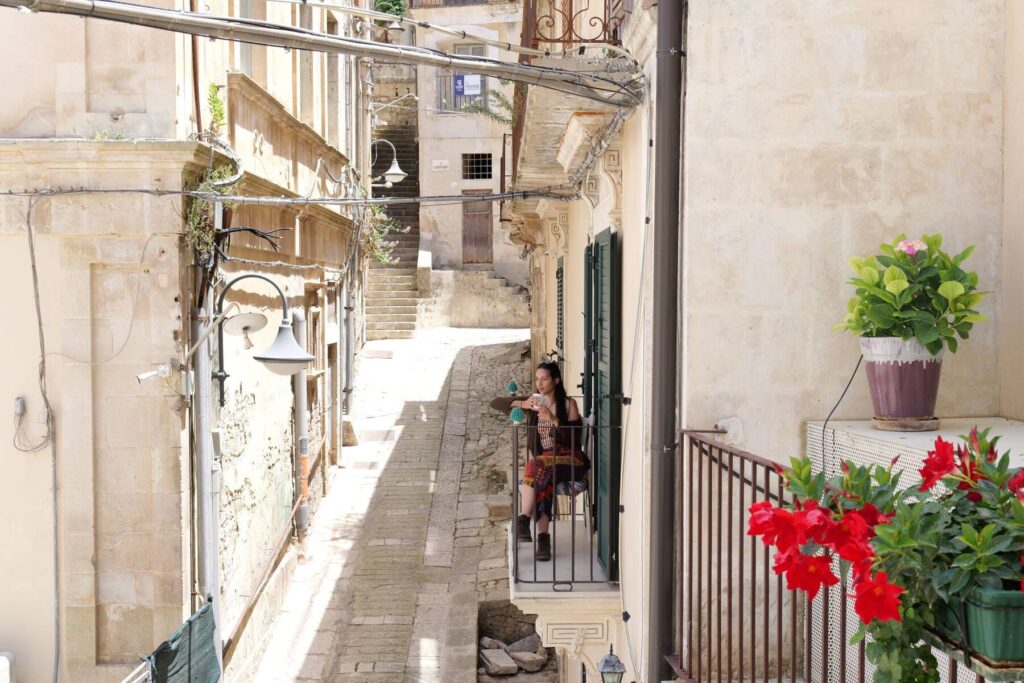 Un'ospite sul balcone affacciato su un vicolo di Modica, tra i gerani
